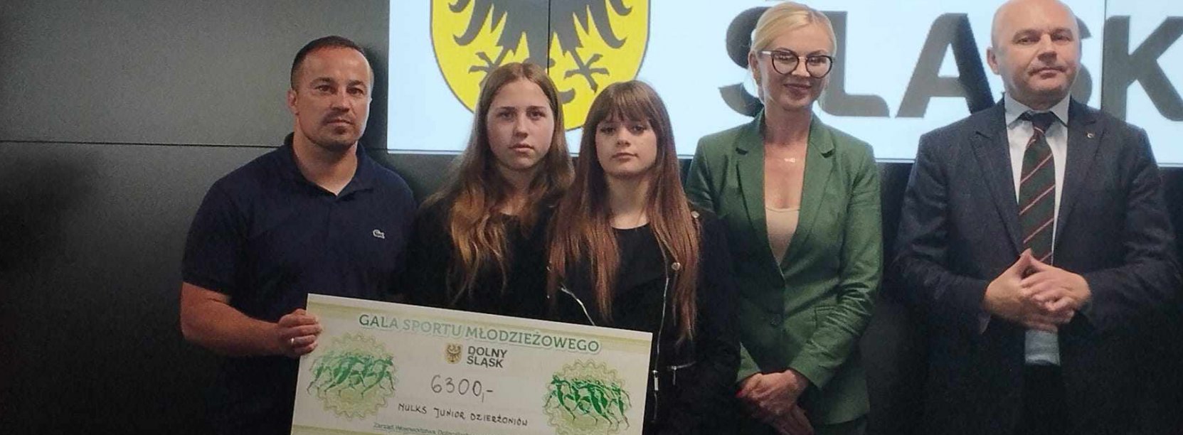 Trener, zawodniczki i zarząd województwa na tla ścianki z napisem Dolny ŚLąsk, w rękach trzrymają czek z nagrodą
