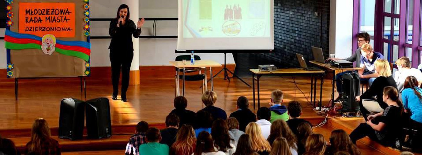 Aula szkolna, dziewczyna stoi przed dużą grupą uczniów i mówi do nich przez mikrofon