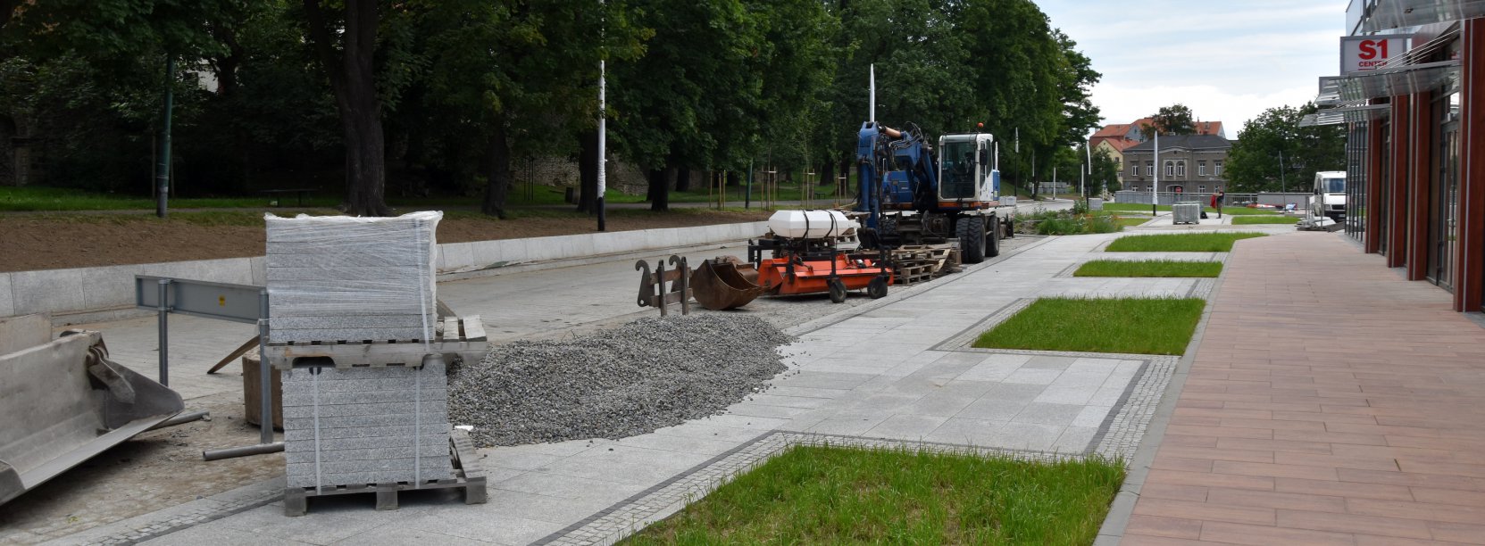 Droga z kostki kamiennej w budowie, obok małe skwery zieleni, na drodze maszyny budowlane