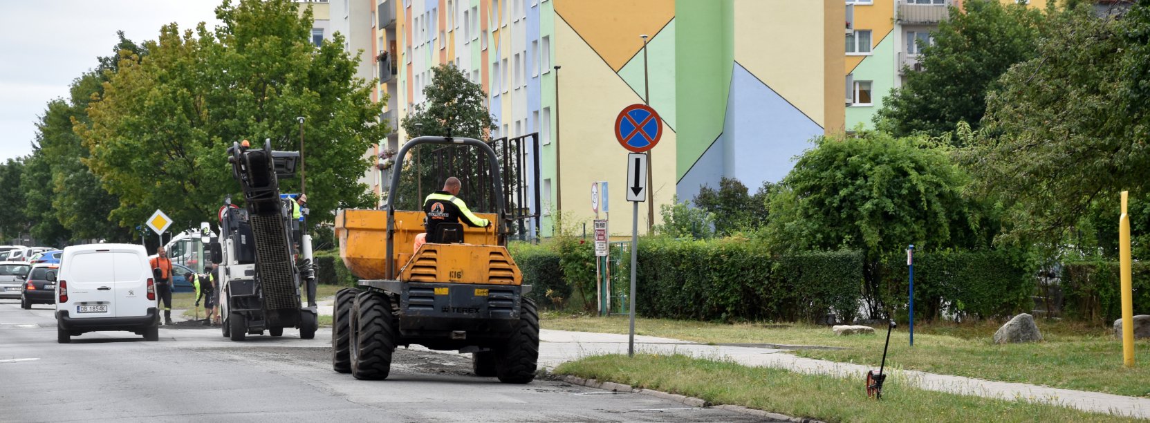 Droga osiedlowa, na części jezdni maszyny do prac drogowych w tle kolorowy blok