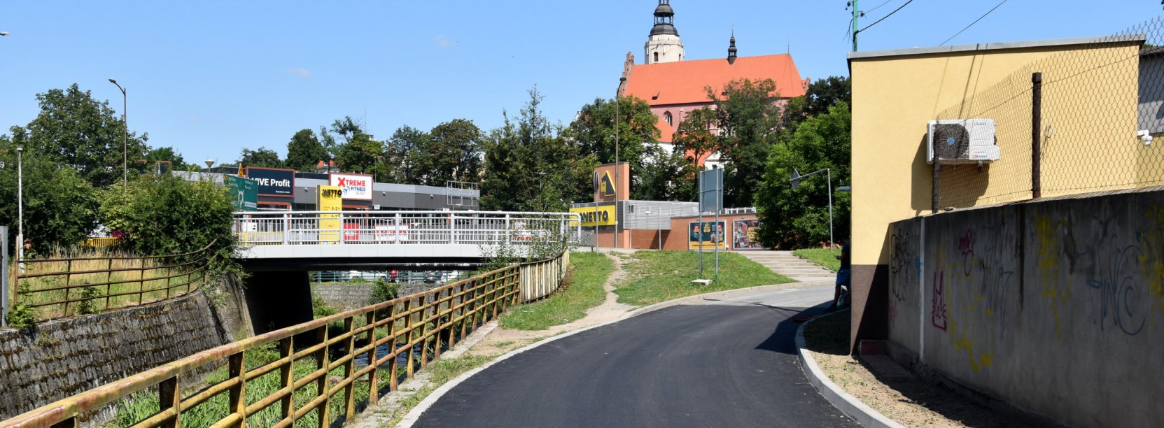Droga z nowym asfaltem, po lewej kamienne koryto rzeki, po prawej mur, w drugim planie średniowieczny kościół