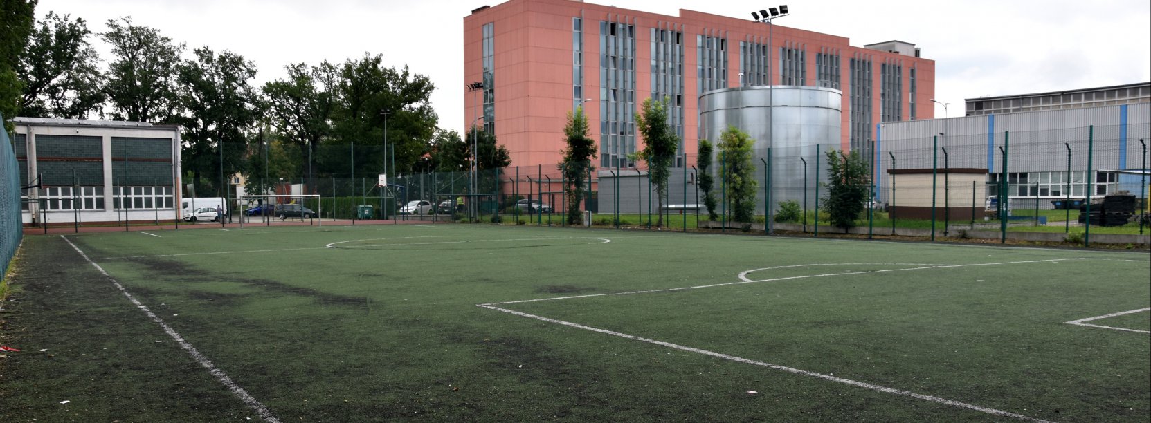 Boisko piłkarskie ze sztuczną trawą, za boiskiem duży czerwony budnek