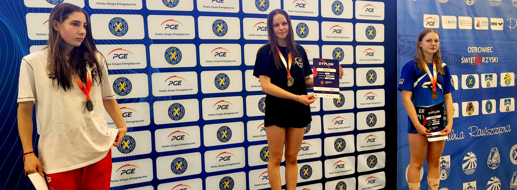 Trzy dziewczyny stojące na podium, za nimi ścianka reklamowa