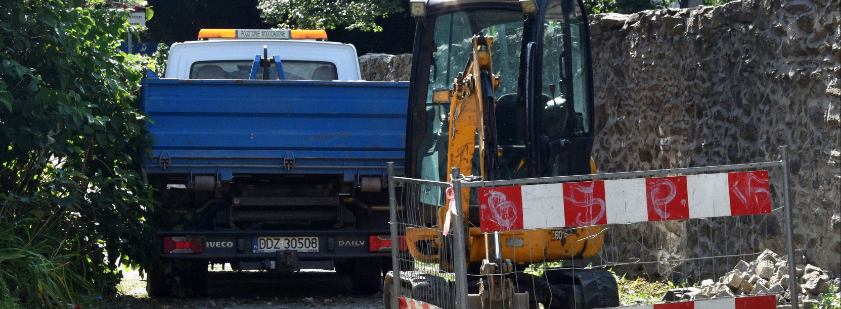 Mała koparka i mały samochód dostawczy na wąskiej drodze, obok drogi kamienny mur