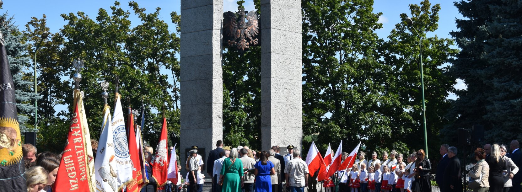 Grupa ludzi stojąca przed pomnikiem, obok poczet sztandarowy i młodzież w białych koszulach 