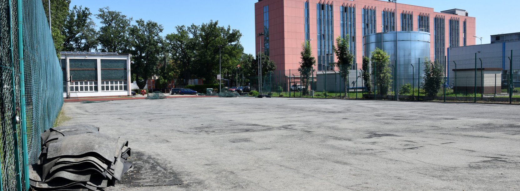 Boisko do piłki nożnej ze zdjętą sztuczną nawierzchnią, w drugim planie czerwony wielopiętrowy budynek