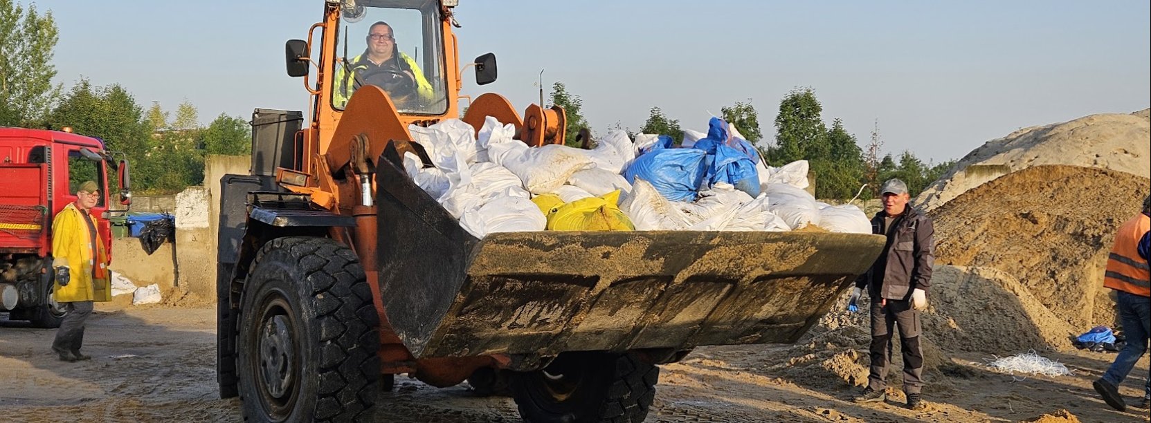 Ładowarka wiezie na "łyżce" worki z piaskiem, w tle cieżąrówka, ludzie i góry piachu