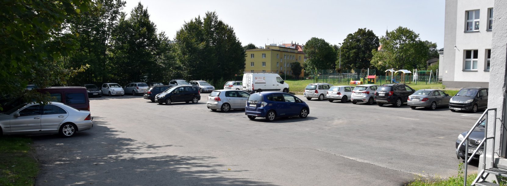 Asfaltowy parking z zaparkowanymi autami, po prawej stronie budynki