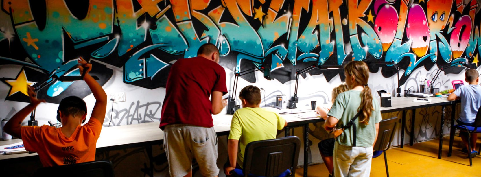 Na tle kolorowej ściany graffiti dzieci siedzą przy stołach i rysują, obok stoi instruktor