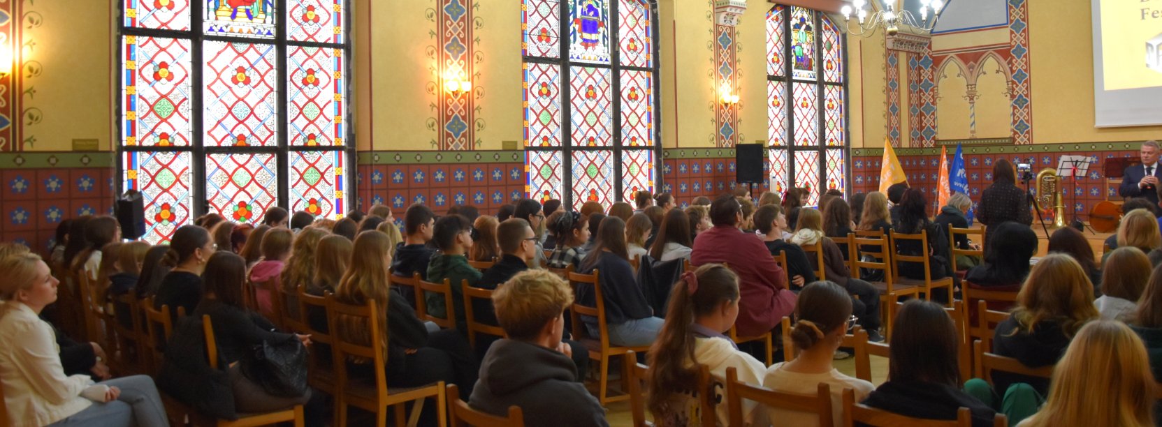 Aula szkolna z dużymi okiennymi witrażami zapełniona uczniami