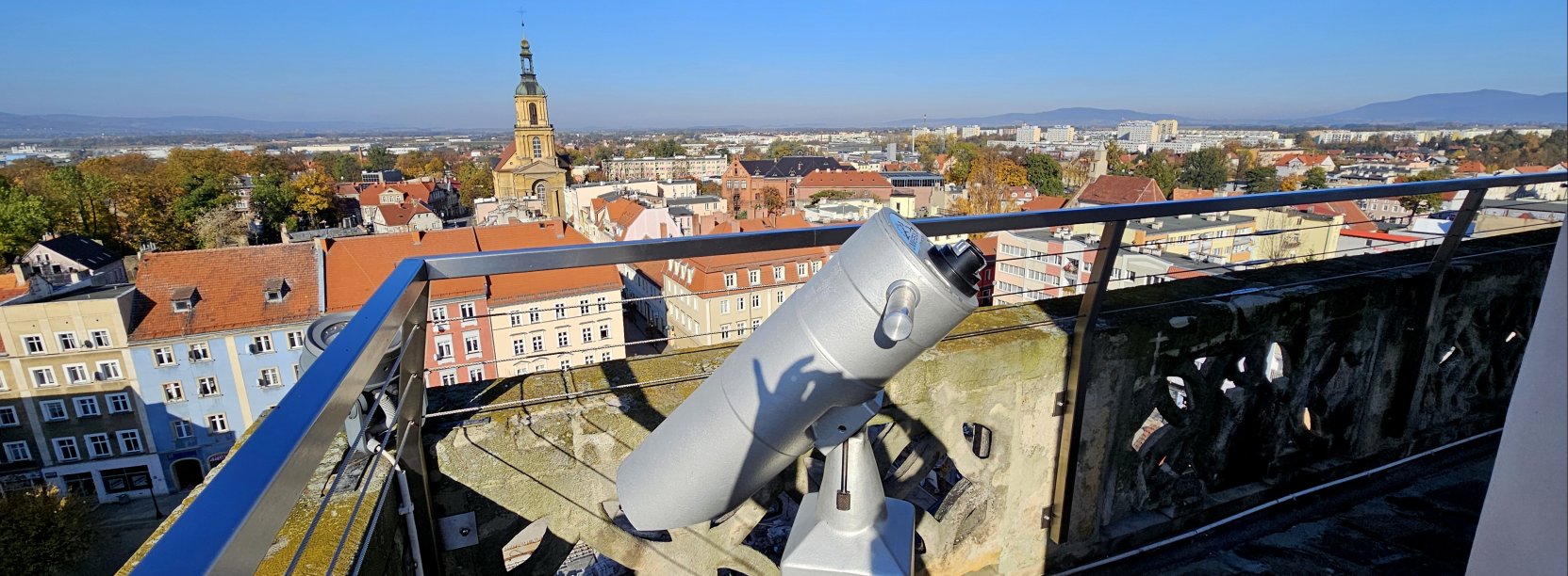 Wieża ratuszowa z kaminnymi i metalowymi barierkami, na pierwszym planie luneta, w tle panorama miasta