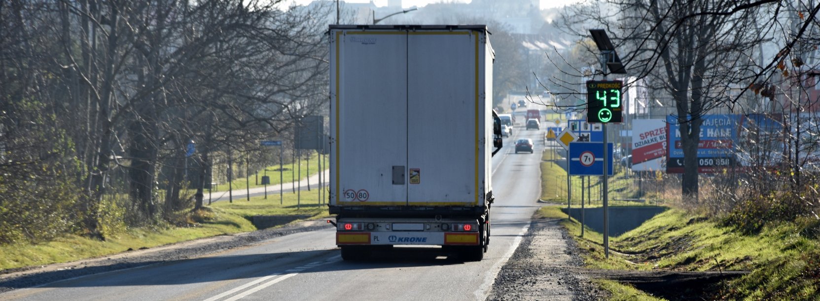 Ciężarówka jadąca drogą wjazdową do Dzierżoniowa, na poboczu tablica informująca kierowców o prędkości