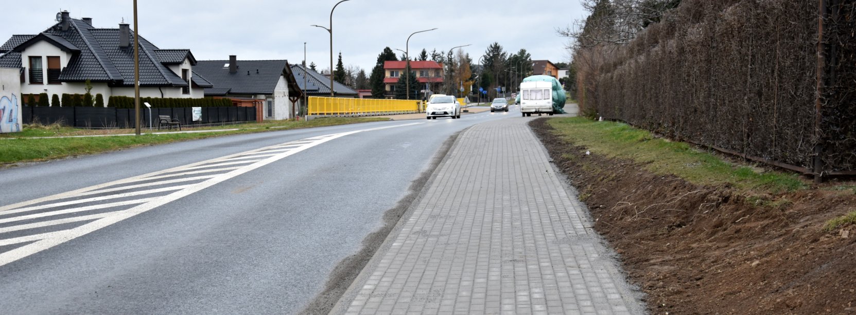 Droga i chodnik z kostki betonowej, po prawej stronie żywopłot, po lewej dom jednorodzinny