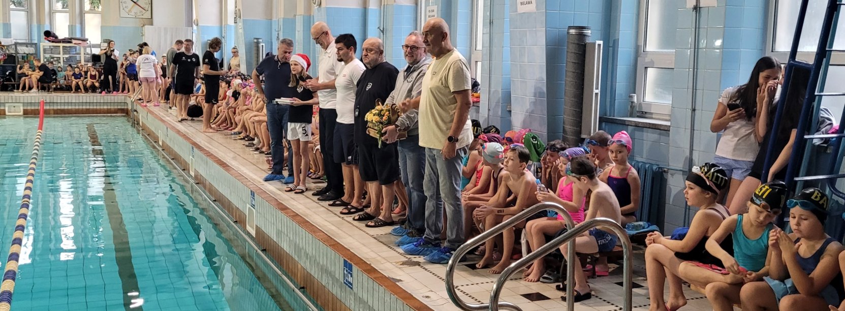 Dzieci siedzące przy krawędzi basenu krytego przed zawodami sportowymi
