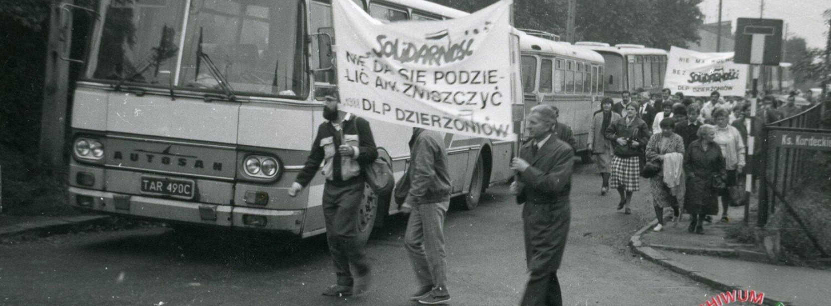 Archwialne zdjęcie ze stanu wojennego, ludzie idący w kolumnie, niosący napisy Solidarność, w tle kolumna autobusów