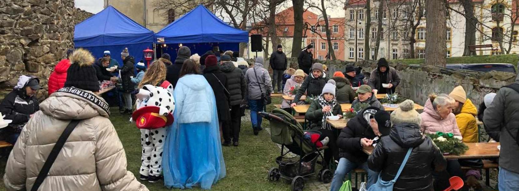 Ludzie siedzący przy stołach, po bokach kamienne, zabytkowe mury obronne