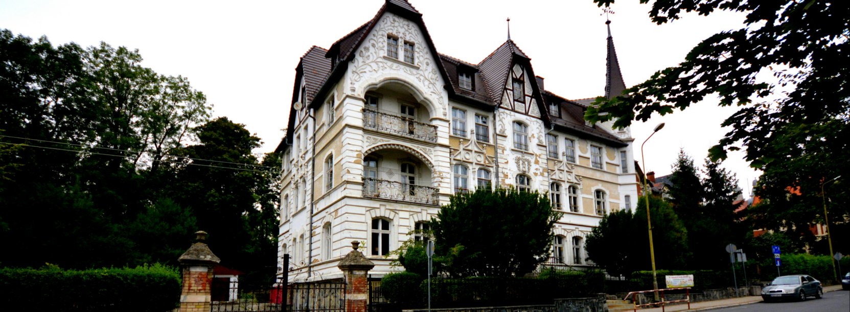 Zabytkowa kamienica ze strzelistą wieżyczką, po bokach duże drzewa