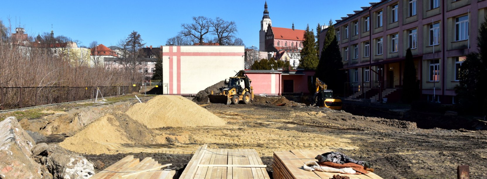 Plac budowy z materiałami budowlanymi, obok trzypiętrowy budynek szkoły