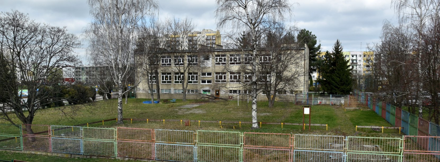 Ogrodzony, duży teren zielony, w tle tzypiętrowy budynek z szarą elewacją