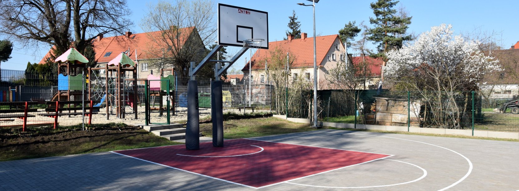 Boisko z kostki betonowej do gry w koszykówkę, za boiskiem plac zabaw i budynki