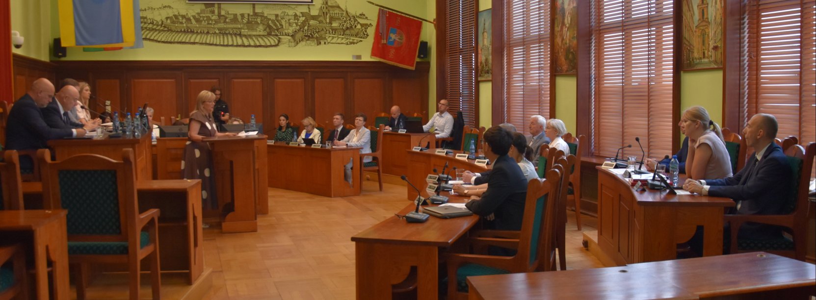 Duża sala z parkietem i drewnianymi stołami, przy stołąch siedzą radni