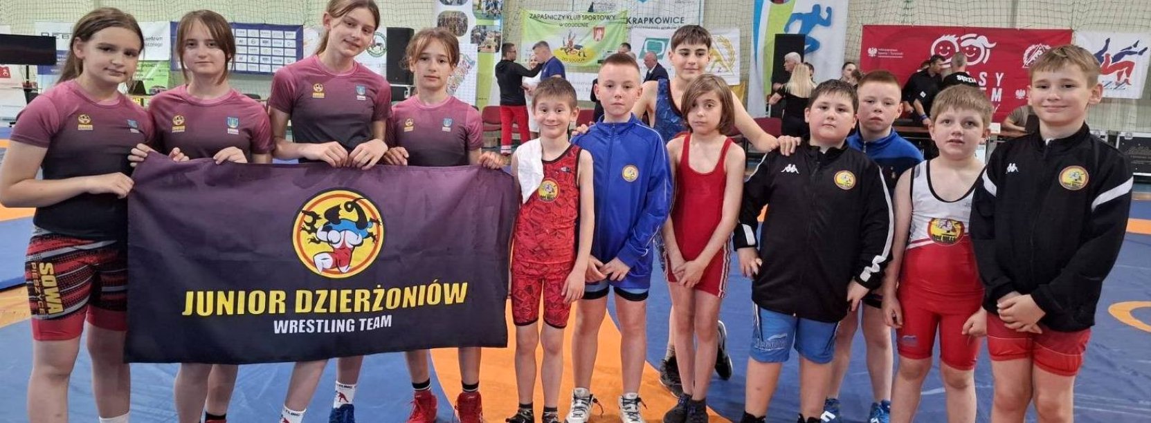 Grupowe zdjęćie kilkunastu młodych zapaśniczek i zapaśników w hali sportowej