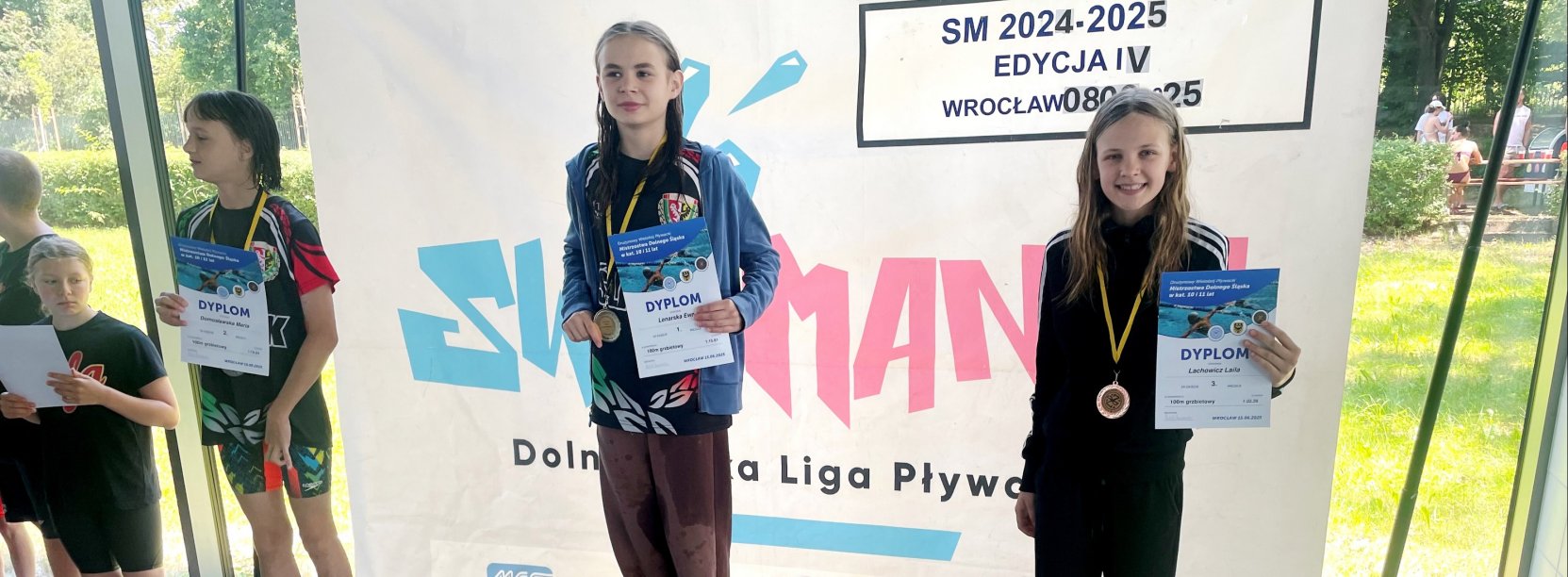 Laila Lachowicz na najwyższym stopniu podium z medalem i dyplomem, po bokach dwie pływaczki