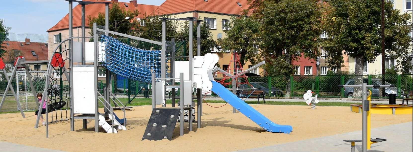 Plac z urządzeniami do zabawy zamontowanymi na piaszczystym podłożu, w tle budynki wielorodzinne