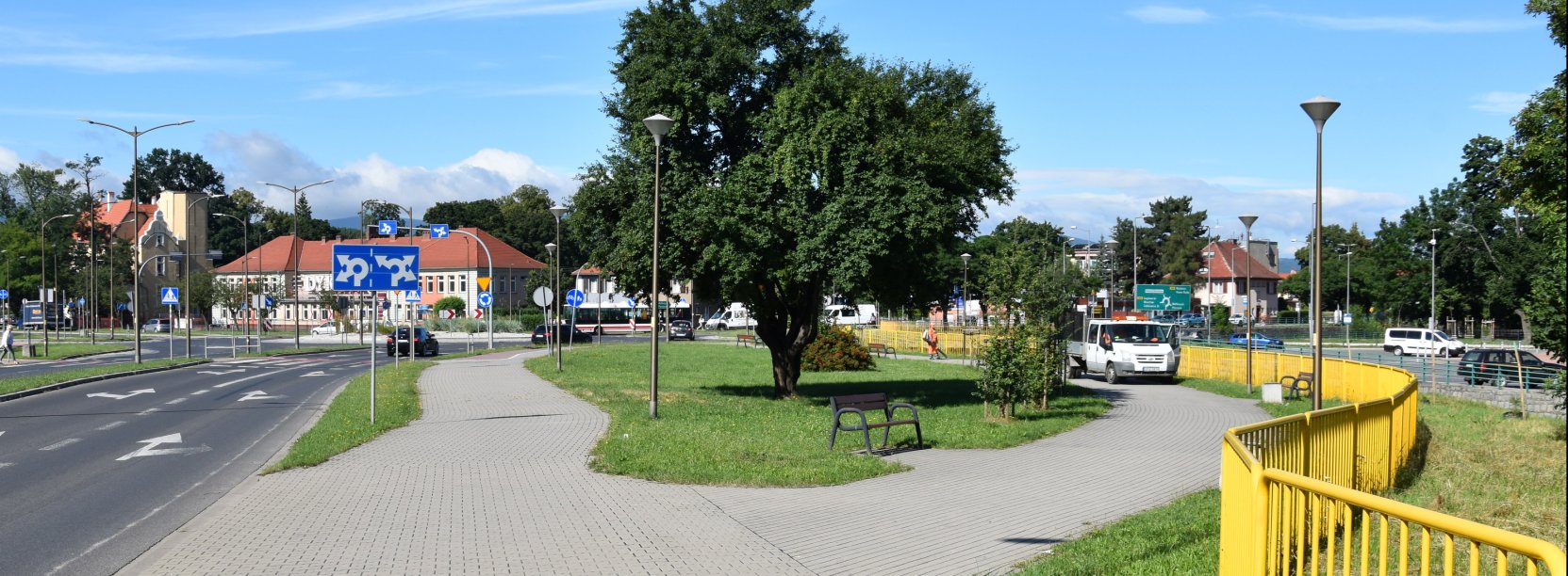Droga w mieście, obok chodnik i teren zielony z dużym drzewem, po prawej żółte barierki