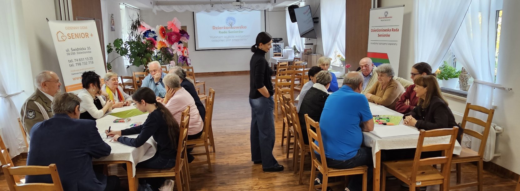 Dwie grupy ludzi przy stołach w sali multimedialnej w Centrum Seniora, pośrodku prowadząca warsztaty pani psycholog