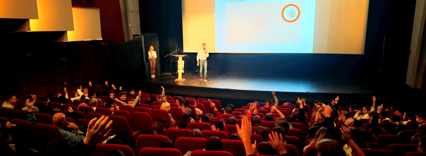 Sala Kinoteatru "Zbyszek", na scenie prowadzący wykład, nad fotelami kinowymi widać ręce zgłaszających się dzieci