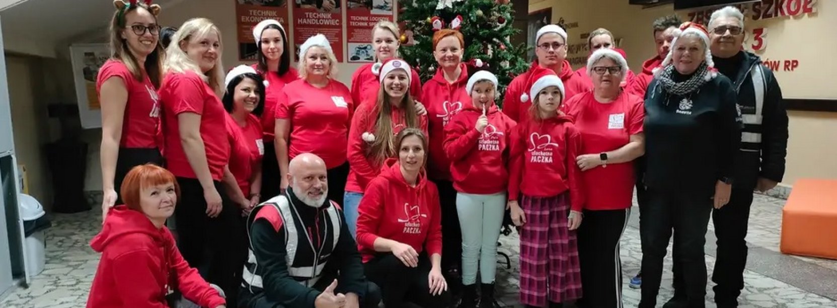 Grupa wolontariuszy Szlechetniej paczki w czerwonych koszulkach i czapkach św. Mikołaja stoi w korytarzu Zespołu Szkół nr 3 w Dzierżoniowie