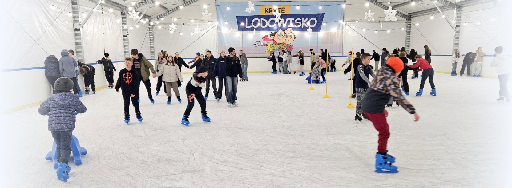 Wnętrze namiotu z lodowiskiem, na tafli lodowiska łyżwiarze w różnym wieku
