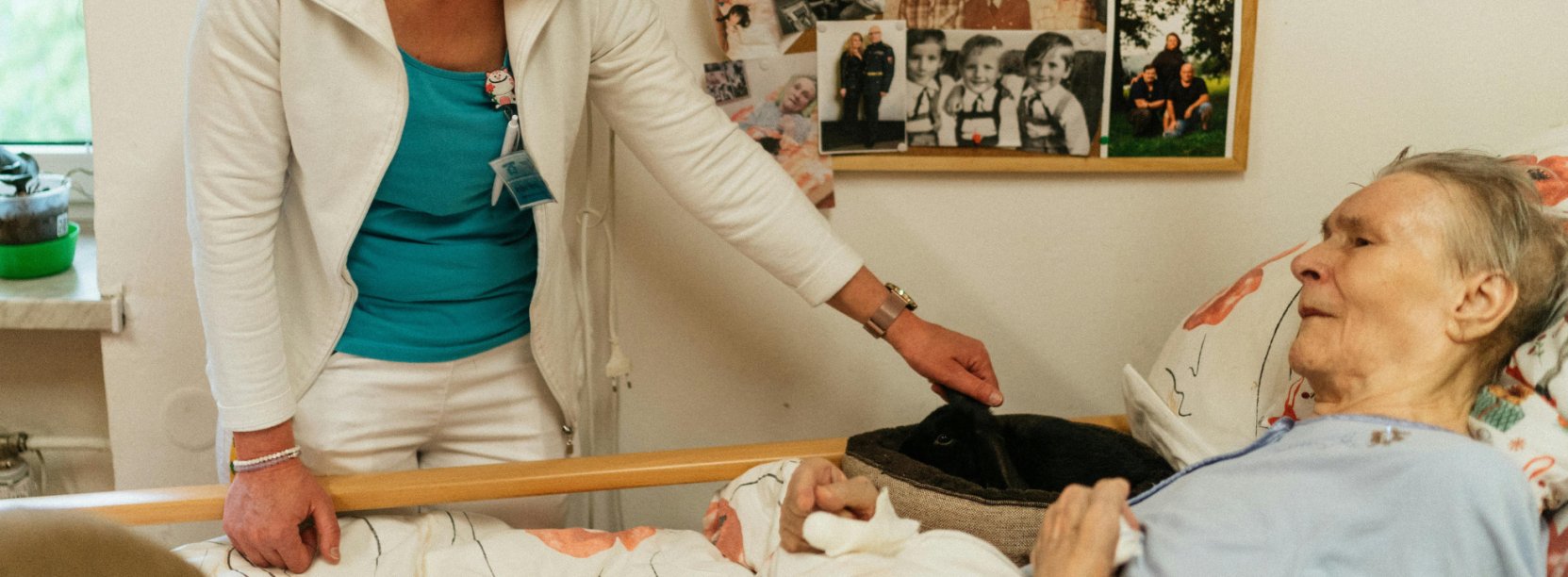 Starsza kobieta leżąca w łóżku, młodsza kobieta stojąca obok się nią opiekuje, na ścianie widoczne fotografie rodzinne