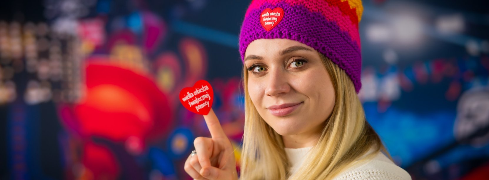 Blondynka w kolorowej czapce s zerduszkiem WOŚP trzyma na palcu serduszko WOŚP