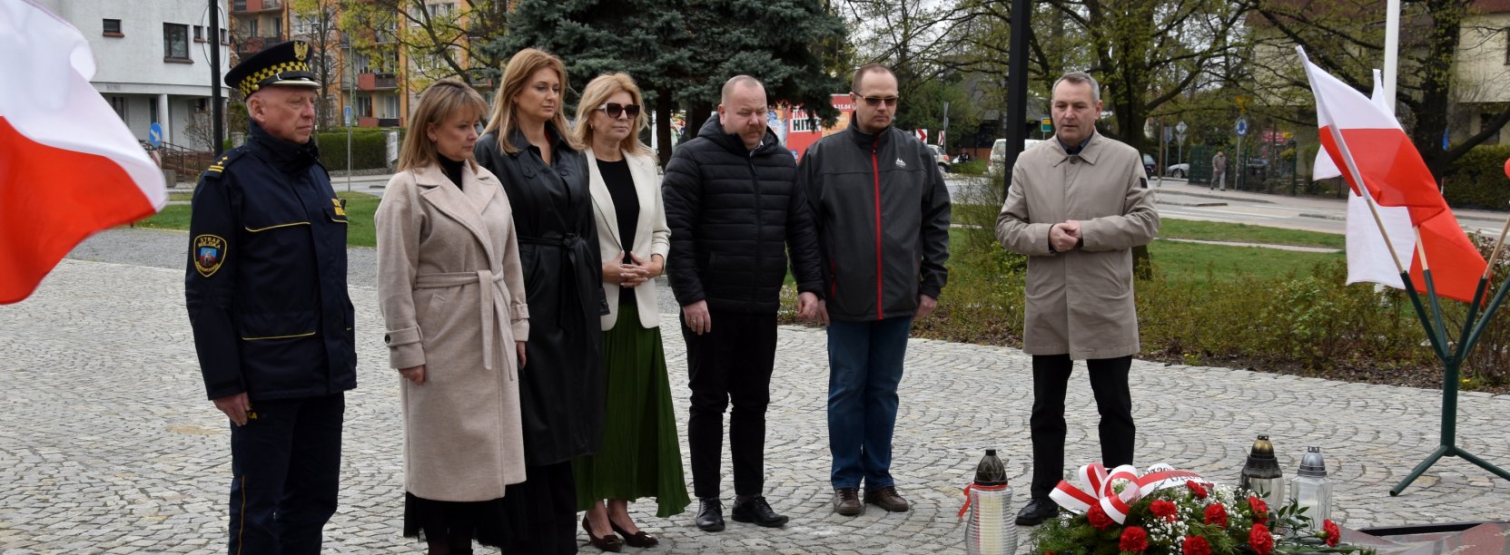 Trzy kobiety i czwórka mężczyzn stoją przed pomnikiem, po bokach flagi Polski