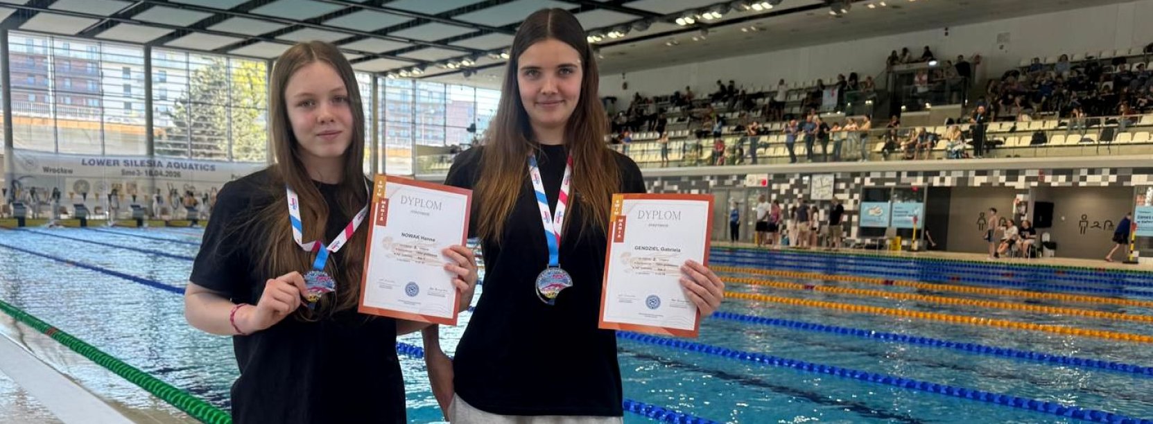 Dwie dziewczyny z medalami i dyplomami stoją przy krawędzi basenu