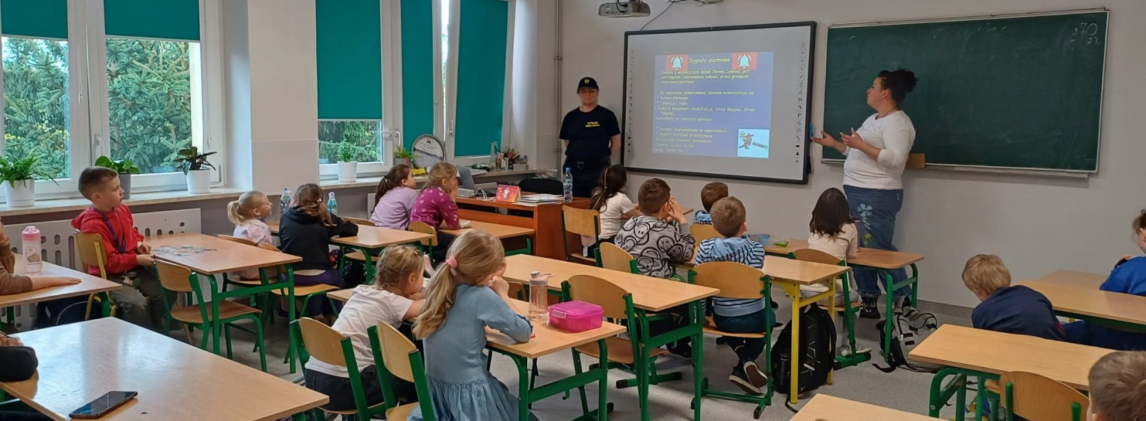 Sala w szkole, dzieci siedzą w ławkach, patrzą w kierunku tablicy, przy niej dwie kobiety, jedna z nich wskazuje ręką na tablicę