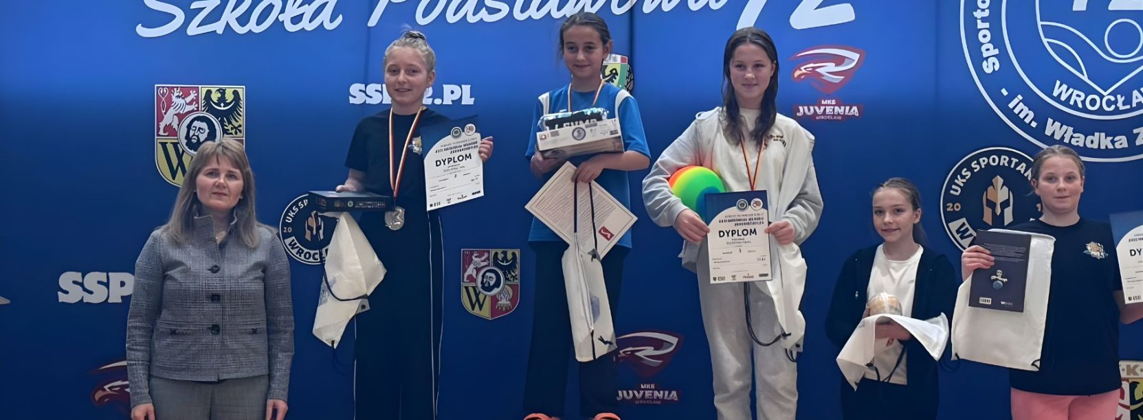 Pięć zawodniczek, trzy stoją na podium, dwie obok, niebieskie tło