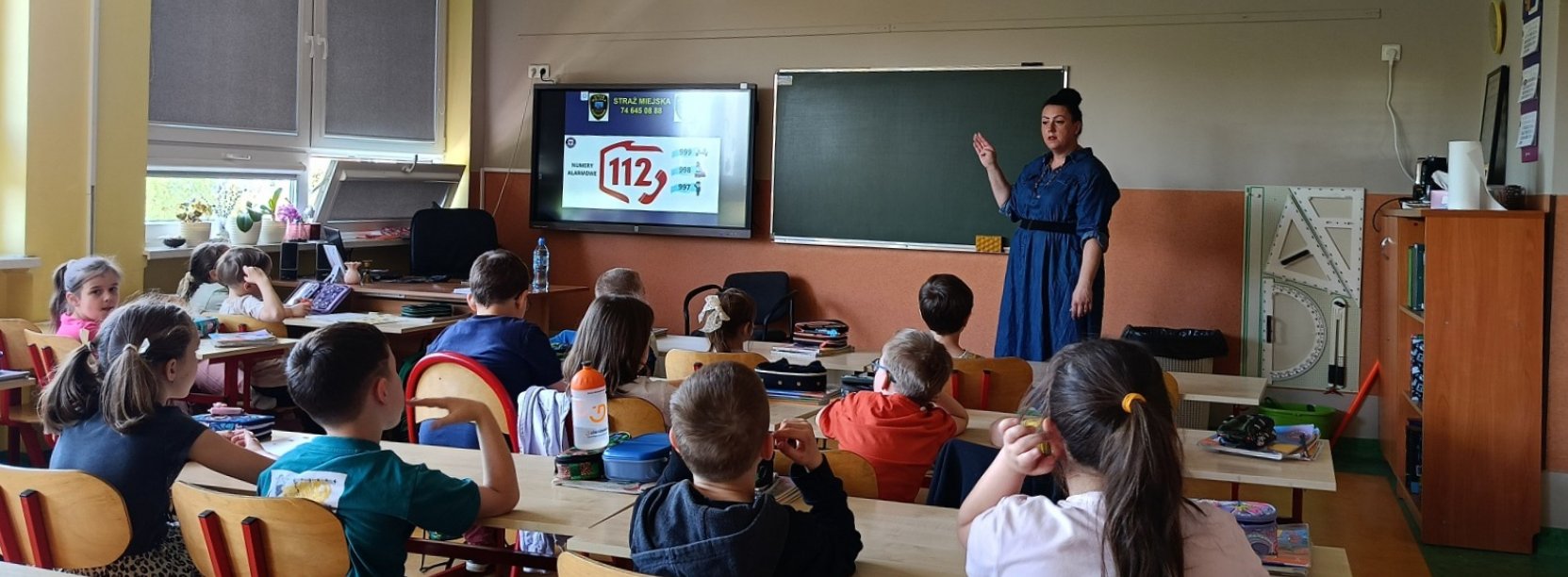 Klasa szkolna, młodzi uczniowie przy ławkach, przy tablicy pani w niebieskiej sukience