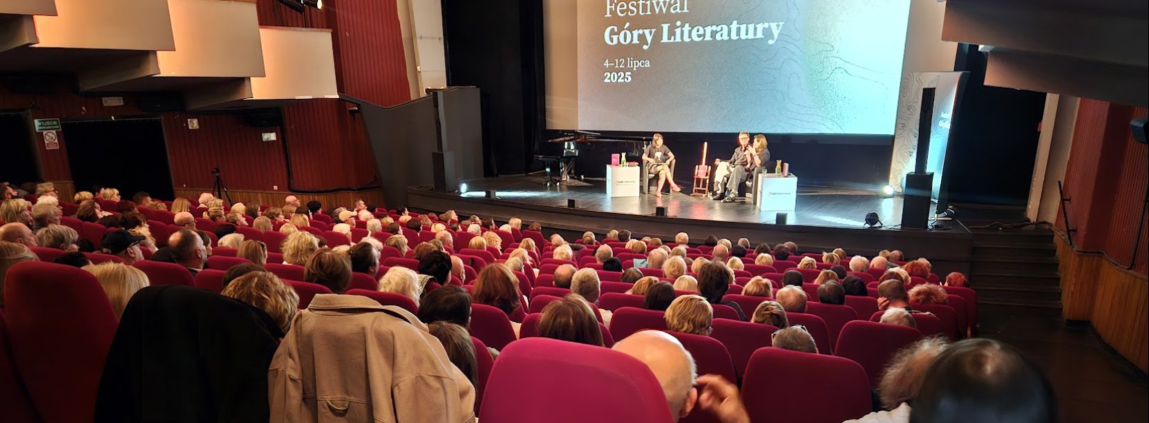 Wypełniona widownia kinoteatru, na scenie dyskusja trzech osób, za nimi na dużym ekranie napis Góry Literatury