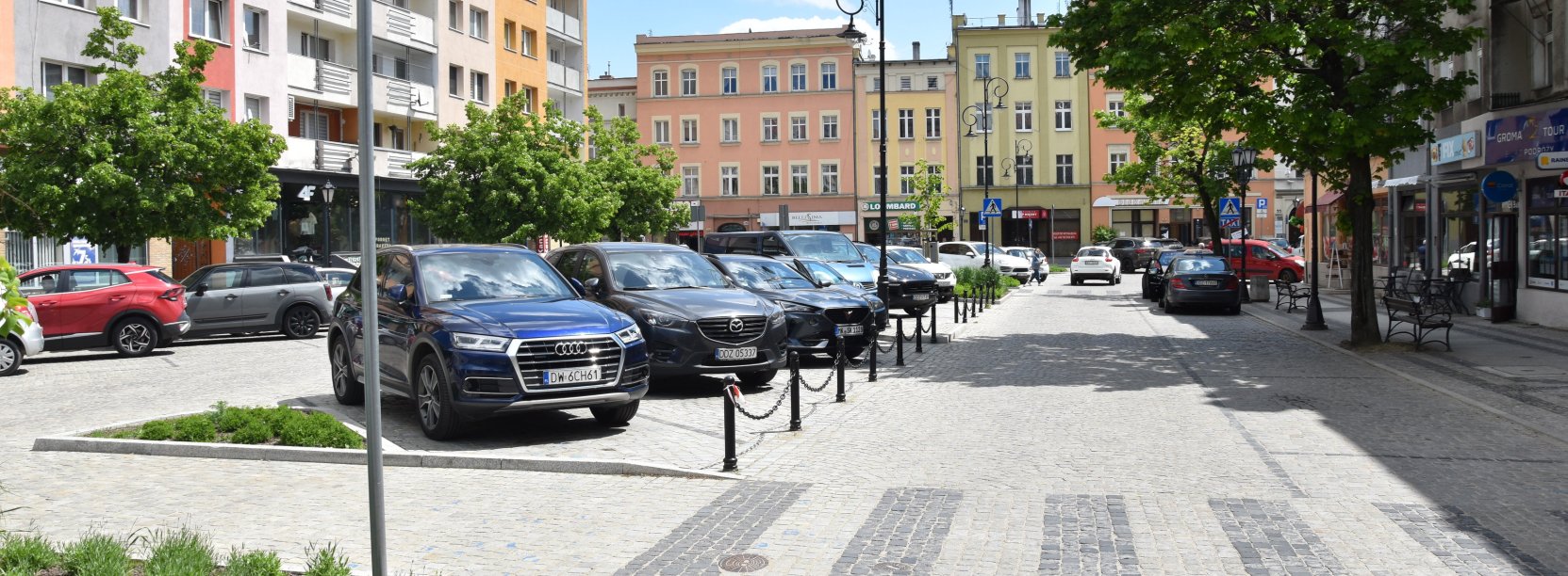 Rynek, zaparkowane samochody, w tle budynki, na pierwszym planie przejście dla pieszych