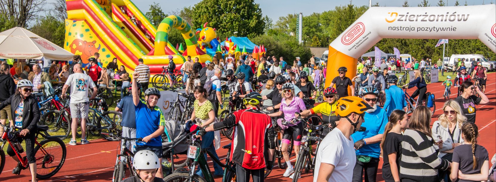 Kilkudziesięciu rowerzystów na bieżni i terenie zielonym, w tle meta i dmuchany zamek dla dzieci