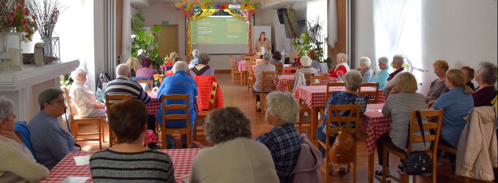Duża sala, przy stołach siedzą seniorzy, na końcu sali kobieta przy dużym ekranie