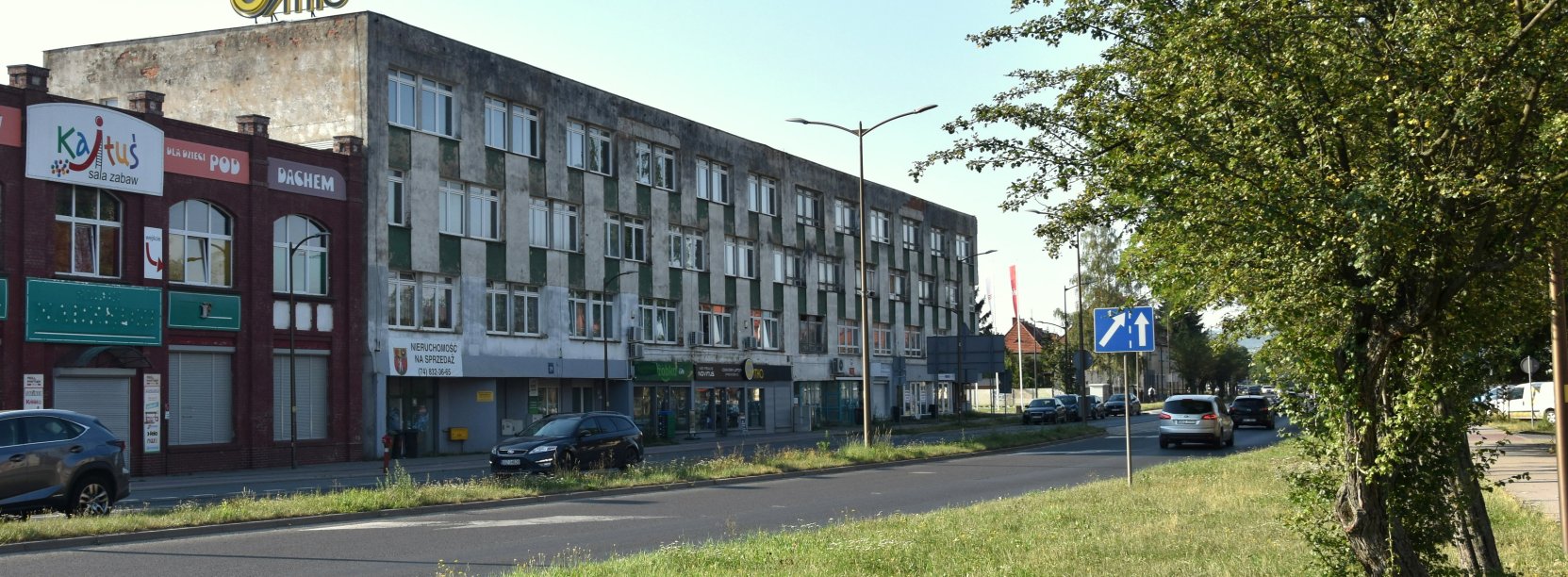 Długi trzypiętrowy budynek położony przy drodze, po prawej stronie drzewo