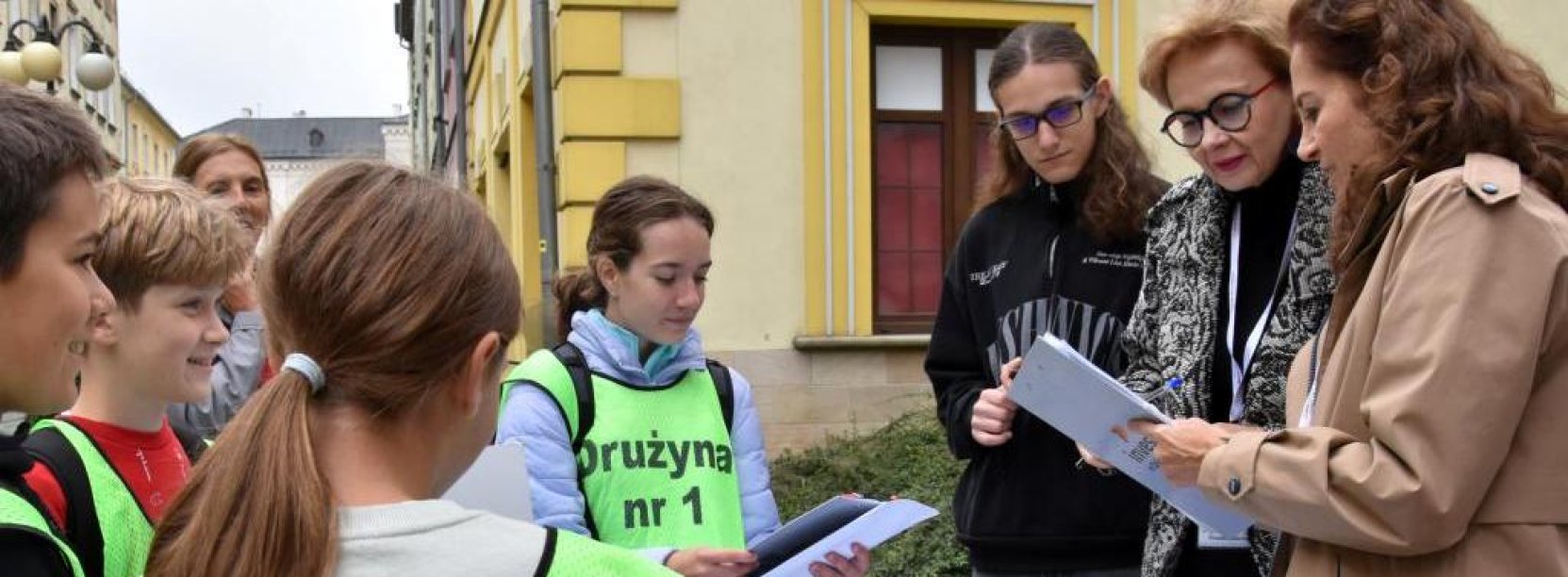 Uczniowie w zielonych kamizelkach z napisem "Gra terenowa" przy stolu ustawionym na ulicy, obok trójka organizatorów konkursu