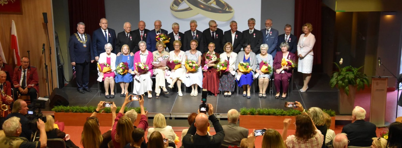 Jubilaci na wspólnym zdjęciu, kobiety siedzą na krzesłach, za nimi stoją mężowie. Przed sceną grupa osób robiących zdjęcia