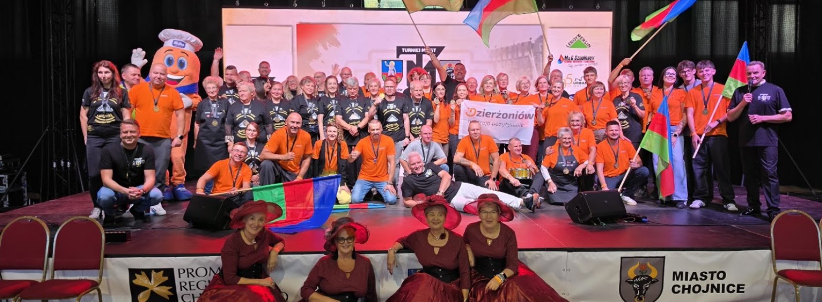 duża grupa osób na scenia w czarnych i pomarańczowych koszulkach, na pierwszym planie cztery panie w bordowych sukniach i kapeluszacj