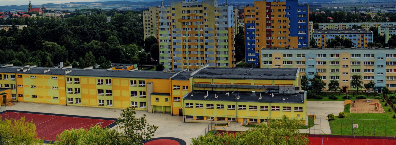 Trzypiętrowy budynek szkoły z żółtą elewacją, za nim wieżowce na tle gór