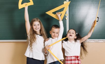 troje dzieci z przyrządami do geometrii stoi przed tablicą w klasie